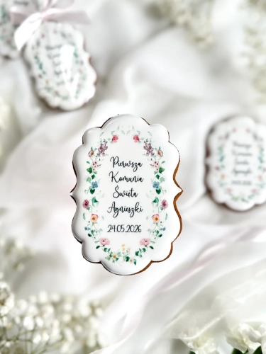 Piernik na komunię w kształcie plakietki z grafiką polnych kwiatów i personalizowanym napisem zapakowany w celofanowy woreczek z kokardką jako forma podziękowania dla gości komunijnych