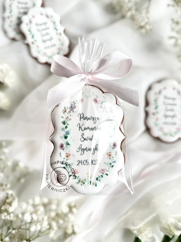 Piernik na komunię w kształcie plakietki z grafiką polnych kwiatów i personalizowanym napisem zapakowany w celofanowy woreczek z kokardką jako forma podziękowania dla gości komunijnych