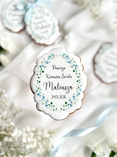 Piernik na komunię w kształcie plakietki z grafiką niebieskich kwiatów i personalizowanym napisem zapakowany w celofanowy woreczek z kokardką jako forma podziękowania dla gości komunijnych