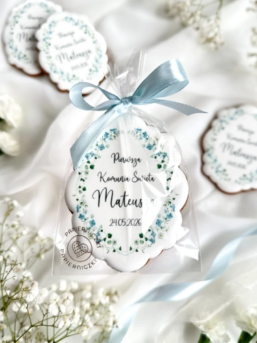 Piernik na komunię w kształcie plakietki z grafiką niebieskich kwiatów i personalizowanym napisem zapakowany w celofanowy woreczek z kokardką jako forma podziękowania dla gości komunijnych