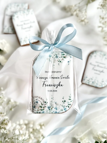 Piernik na komunię w kształcie plakietki z grafiką niebieskich kwiatów, złoceniem i personalizowanym napisem zapakowany w celofanowy woreczek z kokardką jako forma podziękowania dla gości komunijnych