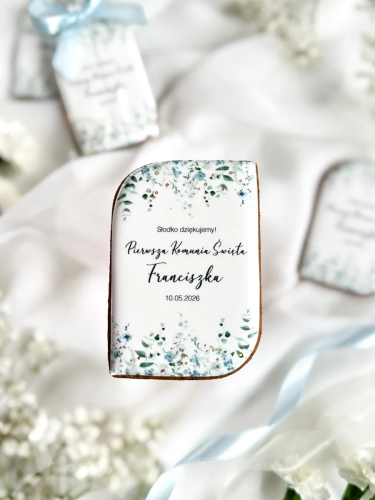 Piernik na komunię w kształcie plakietki z grafiką niebieskich kwiatów, złoceniem i personalizowanym napisem zapakowany w celofanowy woreczek z kokardką jako forma podziękowania dla gości komunijnych