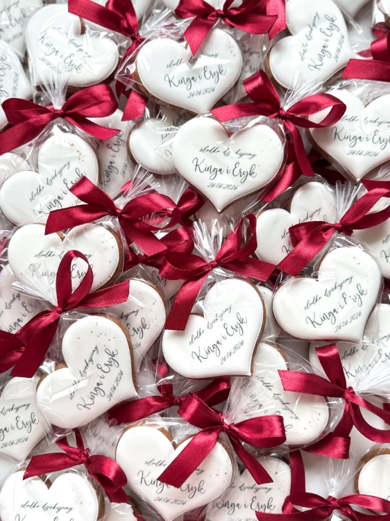 Personalizowane pierniczki w kształcie serca jako podziękowanie dla gości weselnych – białe lukrowane ciasteczka z imionami pary młodej, datą ślubu i bordową kokardą
